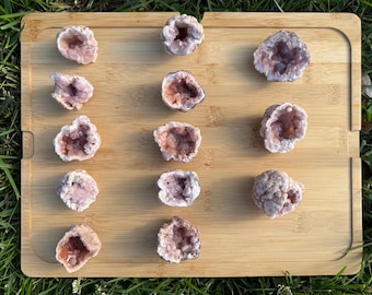 May include: A collection of fourteen small, rough-cut amethyst geode clusters arranged in a grid on a rectangular bamboo board. Each geode features a hollow center filled with light purple to pale pink crystalline structures and a textured exterior.
