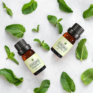 May include: Two amber glass bottles with black caps and white labels containing peppermint essential oil. The bottles are surrounded by fresh green mint leaves on a white background. The labels read "100% Natural & Pure Peppermint Essential Oil Mentha Piperita."