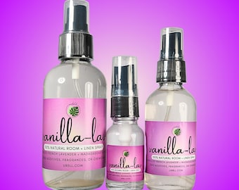 May include: Three clear glass spray bottles of varying sizes, each with a silver cap and a pink label. The labels read "vanilla-lav" and "100% Natural Room + Linen Spray". The background is a gradient of purple.