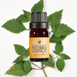 May include: A small amber glass bottle of patchouli essential oil with a black cap, set against a backdrop of fresh green leaves. The label reads "100% Natural & Pure Patchouli Essential Oil". The product is likely used for aromatherapy.