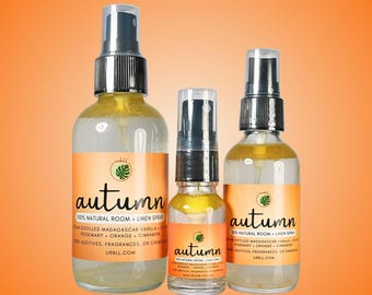 May include: Three glass bottles of room and linen spray in different sizes. The bottles are labeled "autumn" and list the ingredients as "steam distilled Madagascar vanilla, clove, rosemary, orange, cinnamon". The bottles are on a bright orange background.