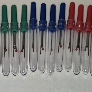 May include: A row of seam rippers in clear plastic tubes. The seam rippers have coloured handles: green, blue, and red. Each ripper has a silver blade and a red safety ball at the tip. These tools are for removing stitches from fabric.