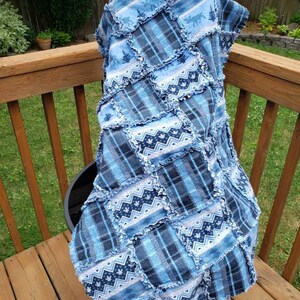 May include: A blue patchwork quilt with a plaid design and forest and geometric patterns. The quilt is displayed over a wooden railing, highlighting its layered construction and fringed edges. The quilt is made of soft fabric.