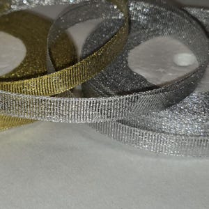 May include: Close-up of shimmering gold and silver ribbon spools. The ribbons have a textured, woven appearance and are arranged in overlapping loops. The metallic sheen suggests a decorative or craft application.