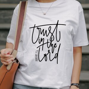 Op de afbeelding: Wit T-shirt met de tekst "Trust in the Lord" in zwart script. Het shirt bevat ook de verwijzing "Proverbs 3:5-6". De persoon draagt een blauwe spijkerbroek en een bruine handtas.