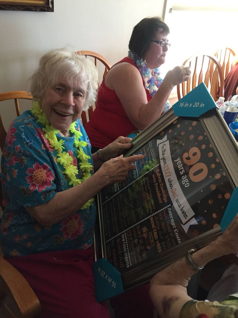 May include: A framed black chalkboard sign with gold glitter accents and white text celebrating a 90th birthday. The sign lists historical events and average costs from the year 1927. The sign is 16 inches by 20 inches.