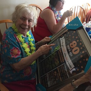 May include: A framed print celebrating a 90th birthday, measuring 16 x 20 inches. The print includes facts from 1927, such as the president and average costs. Two women are holding the framed print, smiling and pointing at it.
