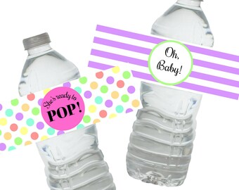 May include: Two water bottle labels for a baby shower. One label has a pink circle with the text "She's ready to POP!" and a colorful polka dot pattern. The other label has a purple and white striped background with a green circle that says "Oh, Baby!"