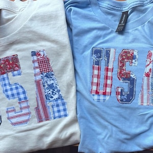 May include: Two crew neck t-shirts, one cream and one light blue, each featuring the letters USA appliqued on the front. The letters are constructed from various fabric patches in red, white, and blue patterns, including gingham, floral, and star prints.
