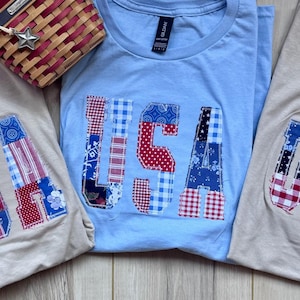 May include: Three short-sleeved crew neck t-shirts, two in beige and one in light blue, each featuring the letters USA appliqued on the front in a patchwork of red, white, and blue patterned fabrics, accompanied by a small woven basket with a star.