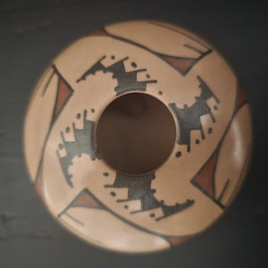May include: A tan-colored ceramic bowl with a central hole and a decorative pattern. The design features black and red geometric shapes arranged around the rim. The bowl has a smooth, rounded shape.