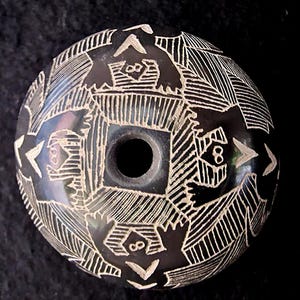 Puede incluir: Una cuenta esférica negra con intrincados diseños blancos grabados. La cuenta presenta patrones geométricos, figuras estilizadas en forma de pájaro y el número "8" dentro de formas cuadradas y triangulares. Se ve un agujero central.