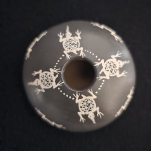 May include: A dark grey, circular object with a central hole, decorated with white, stylised frog designs. The frogs are arranged around the perimeter, with a dotted line pattern. The object has a smooth surface.