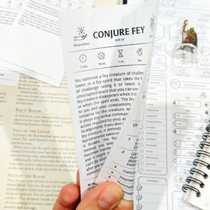 May include: A close-up of a white card with black text. The card is titled "Conjure Fey" and is a 6th level spell. The card describes the spell's effects and how to use it. The card is folded in half and held in a hand.