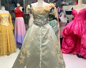 May include: A sage green ball gown with a lace bodice and gold butterfly embroidery. The gown has a fitted bodice and a full skirt.