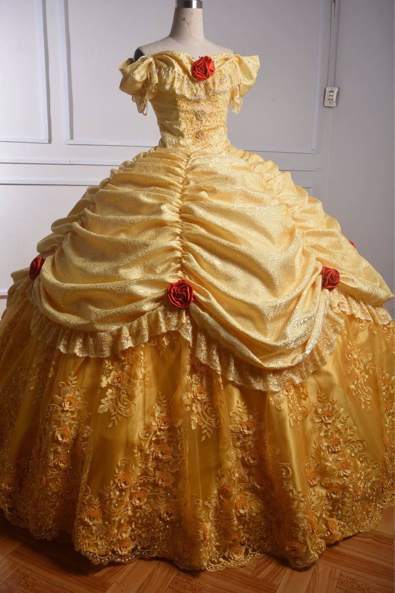 May include: A yellow ball gown with a lace overlay and red rose accents. The dress has a sweetheart neckline and off-the-shoulder sleeves.