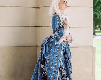 May include: A woman in a blue and white patterned dress with a long train and white lace trim. She is standing in front of a white wall and holding a piece of paper in her hand.