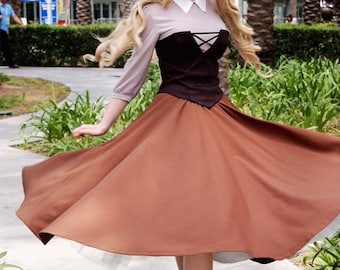 May include: A woman dressed as a princess in a brown and black dress with a white blouse. She has long blonde hair and is wearing a headband. She is standing in front of a green bush and a building.