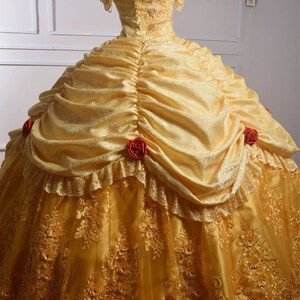 May include: A yellow ball gown with a lace overlay and red rose accents. The dress has a sweetheart neckline and off-the-shoulder sleeves.