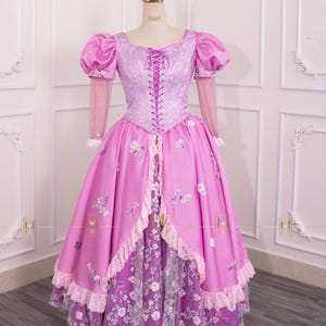 Könnte beinhalten: Ein Prinzessinnenkleid in Rosa und Lila mit Schnürleibchen und Puffärmeln. Das Kleid hat Blumenstickereien und einen geschichteten Rock mit Spitzenbesatz. Eine goldene Krone sitzt auf dem Kopf der Schaufensterpuppe.