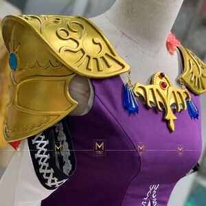 May include: Purple and gold cosplay costume with a gold shoulder piece and a gold necklace with blue and red accents. The costume has a white collar and a white skirt.