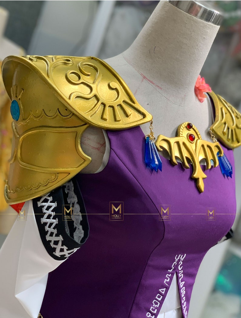 May include: Purple and gold cosplay costume with a gold shoulder piece and a gold necklace with blue and red accents. The costume has a white collar and a white skirt.