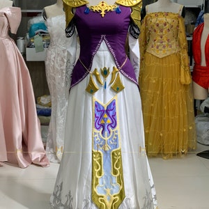 May include: A white and purple princess costume with gold accents and intricate embroidery. The dress features a long skirt, a fitted bodice, and a high neckline. The costume is inspired by a popular video game character.