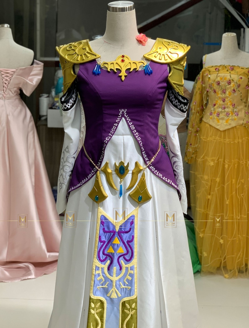 May include: A white and purple dress with gold accents and a blue and green design on the skirt. The dress has a high neckline and long sleeves. The dress is on a mannequin.