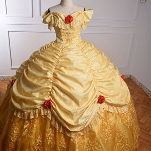 May include: A yellow ball gown with a lace overlay and red rose accents. The dress has a sweetheart neckline and off-the-shoulder sleeves.