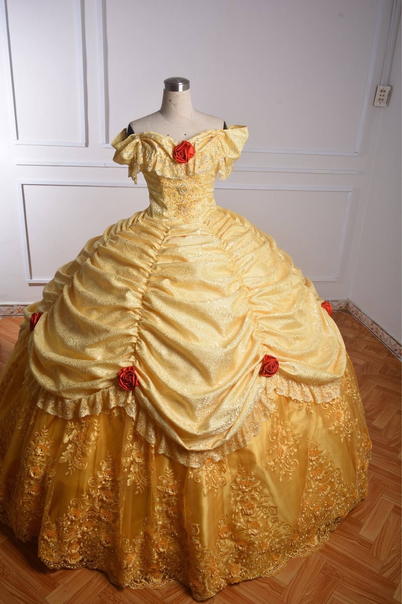 May include: A yellow ball gown with a lace overlay and red rose accents. The dress has a sweetheart neckline and off-the-shoulder sleeves.