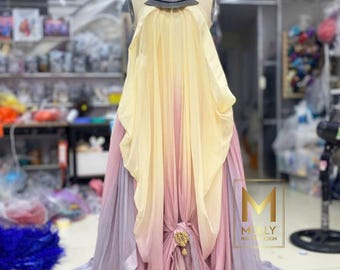 May include: A floor-length gown on a mannequin featuring a layered ombre design transitioning from pale yellow at the top to soft pink and light purple at the hem. The dress includes a dark metallic neck piece and a gold-toned decorative brooch.