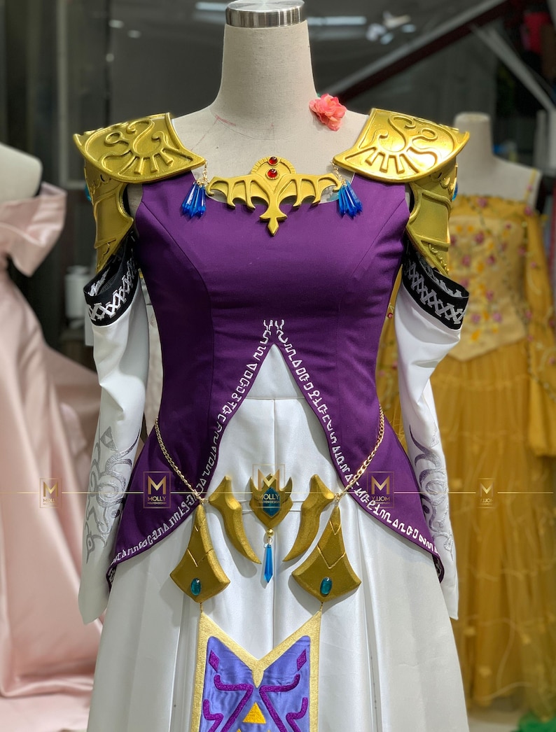 May include: A purple and white dress with gold accents and a gold crown. The dress has a white skirt with a purple and gold design. The dress is on a mannequin.