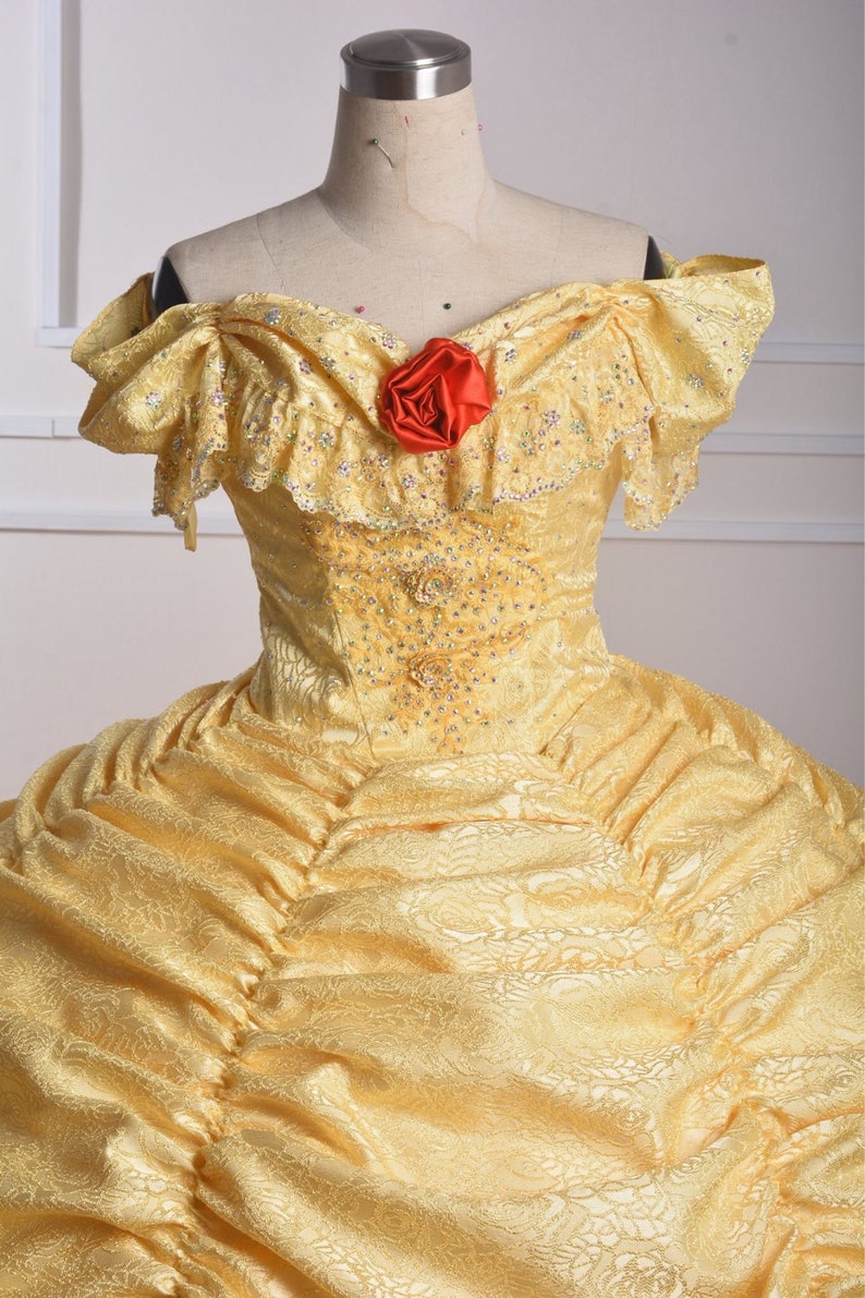 May include: A yellow lace ball gown with a sweetheart neckline and a red rose accent. The gown has a fitted bodice and a full skirt.