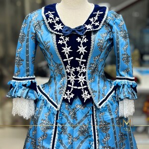 May include: A blue and white dress with a velvet collar and a floral pattern. The dress has a fitted bodice and a full skirt. The dress is decorated with white embroidery and black trim.
