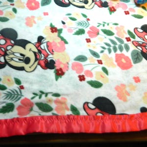 May include: A white blanket with a pattern of Minnie Mouse, floral designs in pink and peach, and green leaves. It has a bright pink satin border. The blanket is suitable for a cot or a bed.