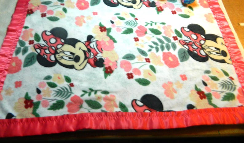 May include: A white blanket with a pattern of Minnie Mouse, floral designs in pink and peach, and green leaves. It has a bright pink satin border. The blanket is suitable for a cot or a bed.