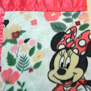 May include: A soft, white fleece blanket featuring a Minnie Mouse graphic with a red polka dot bow and dress. The blanket is decorated with a floral pattern in shades of pink, yellow, and green, and is trimmed with a pink satin border.