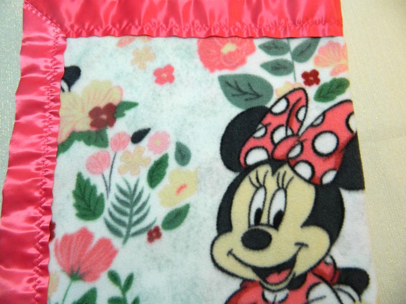 May include: A soft, white fleece blanket featuring a Minnie Mouse graphic with a red polka dot bow and dress. The blanket is decorated with a floral pattern in shades of pink, yellow, and green, and is trimmed with a pink satin border.