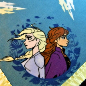 May include: A blue fleece blanket with a yellow satin border. The blanket features a circular design with a cartoon image of Elsa and Anna from the movie Frozen. The characters are facing away from each other, with Elsa on the left and Anna on the right. The background is a blue and white pattern.