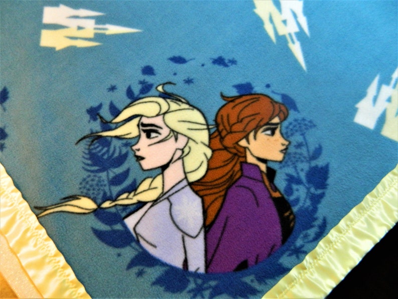 May include: A blue fleece blanket with a yellow satin border. The blanket features a circular design with a cartoon image of Elsa and Anna from the movie Frozen. The characters are facing away from each other, with Elsa on the left and Anna on the right. The background is a blue and white pattern.
