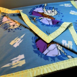 May include: A blue fleece blanket with a yellow satin border. The blanket features a repeating pattern of a white castle and a floral design with two girls from the movie Frozen.
