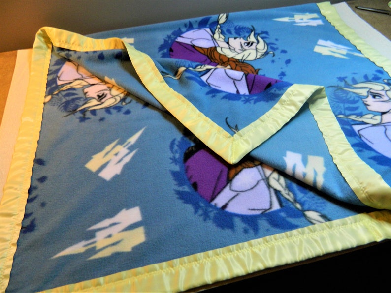 May include: A blue fleece blanket with a yellow satin border. The blanket features a repeating pattern of a white castle and a floral design with two girls from the movie Frozen.