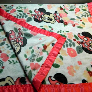 May include: A white fleece blanket with a floral pattern and Minnie Mouse illustrations. The blanket has a pink satin border. The design includes pink, peach, and yellow flowers with green leaves.