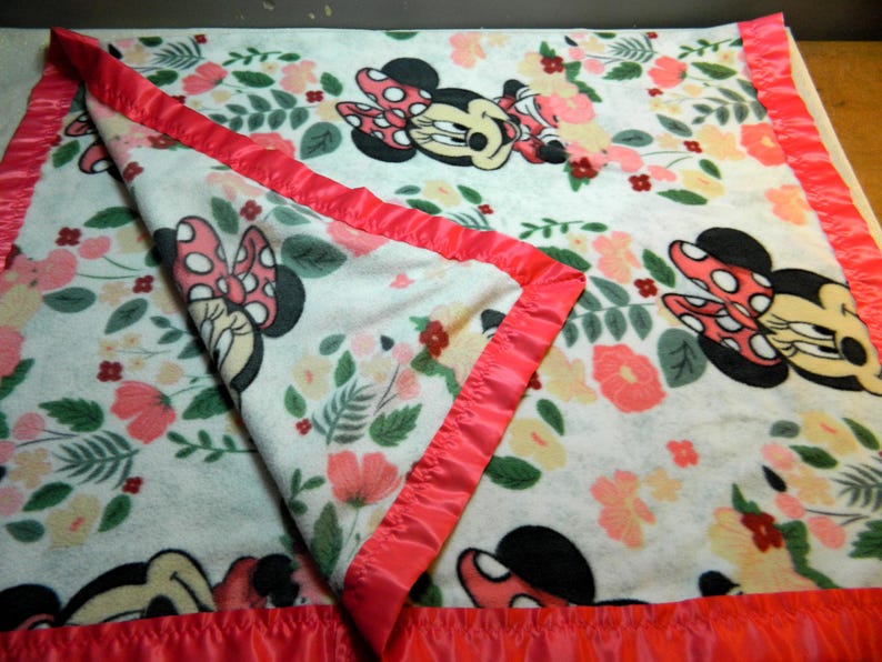 May include: A white fleece blanket with a floral pattern and Minnie Mouse illustrations. The blanket has a pink satin border. The design includes pink, peach, and yellow flowers with green leaves.