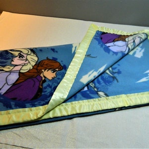 May include: A blue fleece blanket with a yellow satin border. The blanket features a design of two girls from the movie Frozen.
