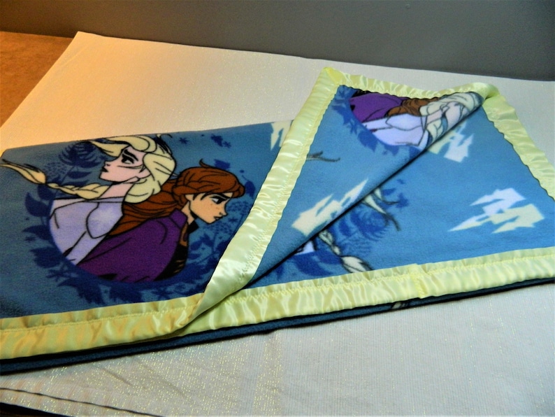 May include: A blue fleece blanket with a yellow satin border. The blanket features a design of two girls from the movie Frozen.