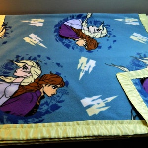 May include: A blue fleece blanket with a satin border. The blanket features a repeating pattern of two girls with long hair, one with blonde hair and one with brown hair. The girls are wearing purple and blue dresses. The background is a light blue with white snowflakes.