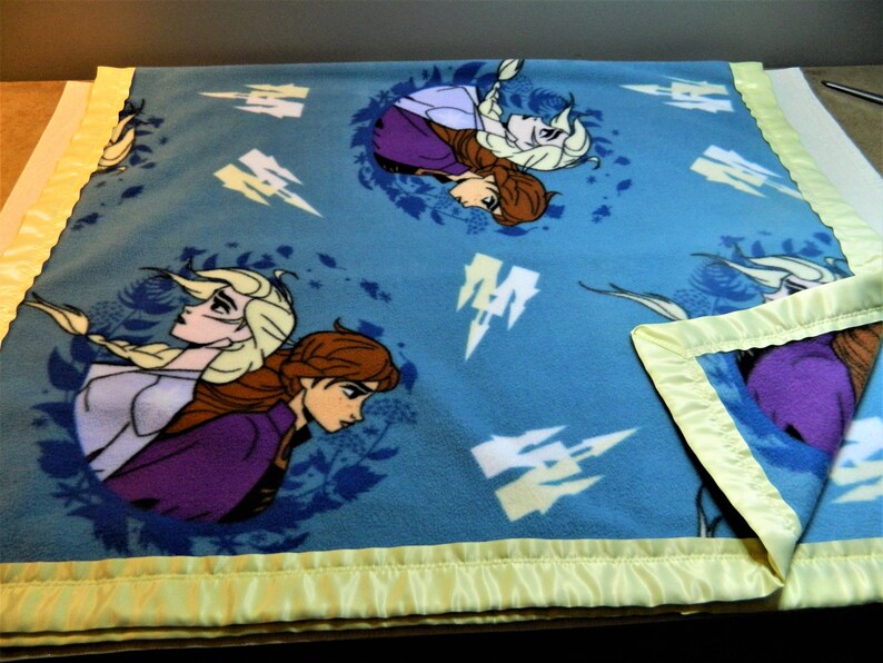 May include: A blue fleece blanket with a satin border. The blanket features a repeating pattern of two girls with long hair, one with blonde hair and one with brown hair. The girls are wearing purple and blue dresses. The background is a light blue with white snowflakes.