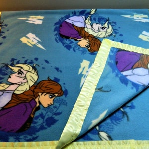 May include: A blue fleece blanket with a yellow satin border. The blanket features a repeated pattern of two girls with long blonde and brown hair. The girls are wearing purple and blue dresses. The background is a light blue with white lightning bolts.