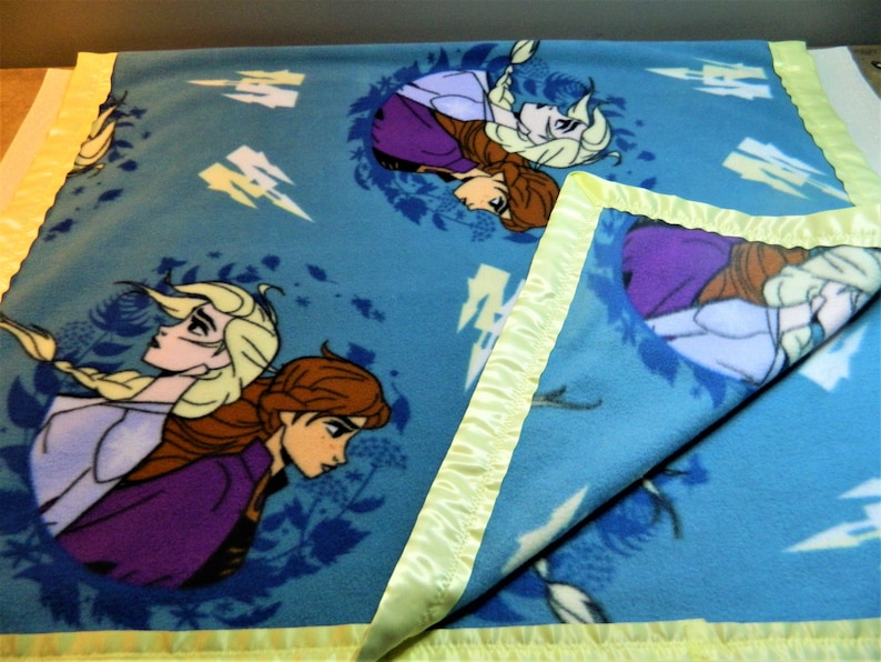 May include: A blue fleece blanket with a yellow satin border. The blanket features a repeated pattern of two girls with long blonde and brown hair. The girls are wearing purple and blue dresses. The background is a light blue with white lightning bolts.